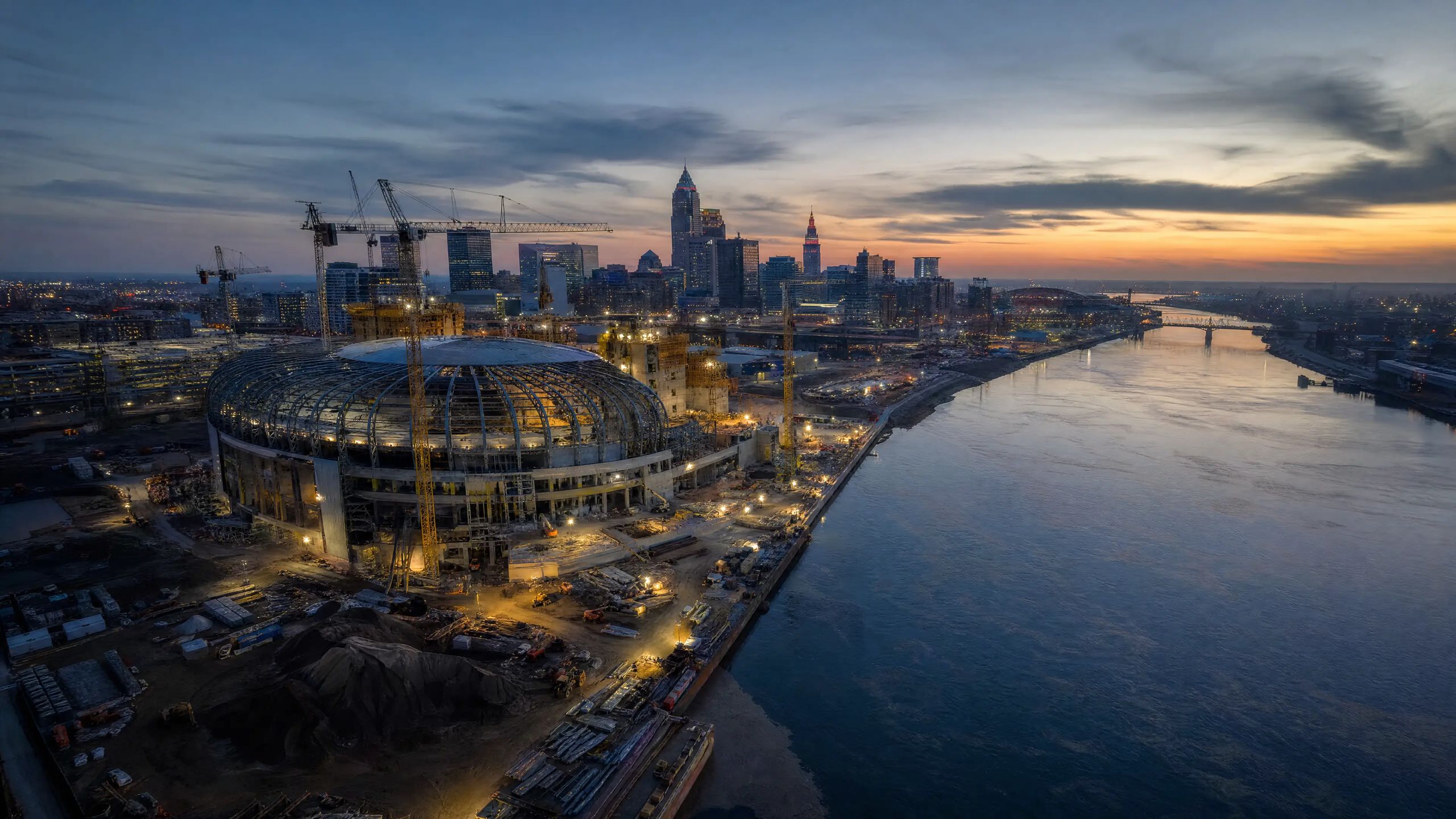 Riverfront construction site with a domed stadium under development at dusk